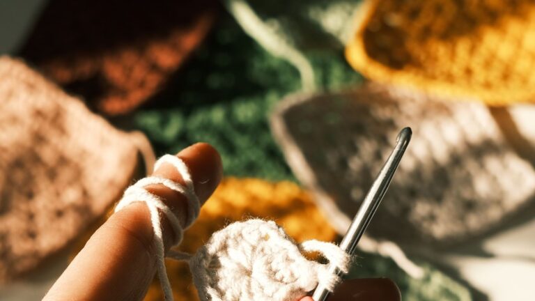 Mãos fazendo crochê com linha branca, quadrados coloridos ao fundo